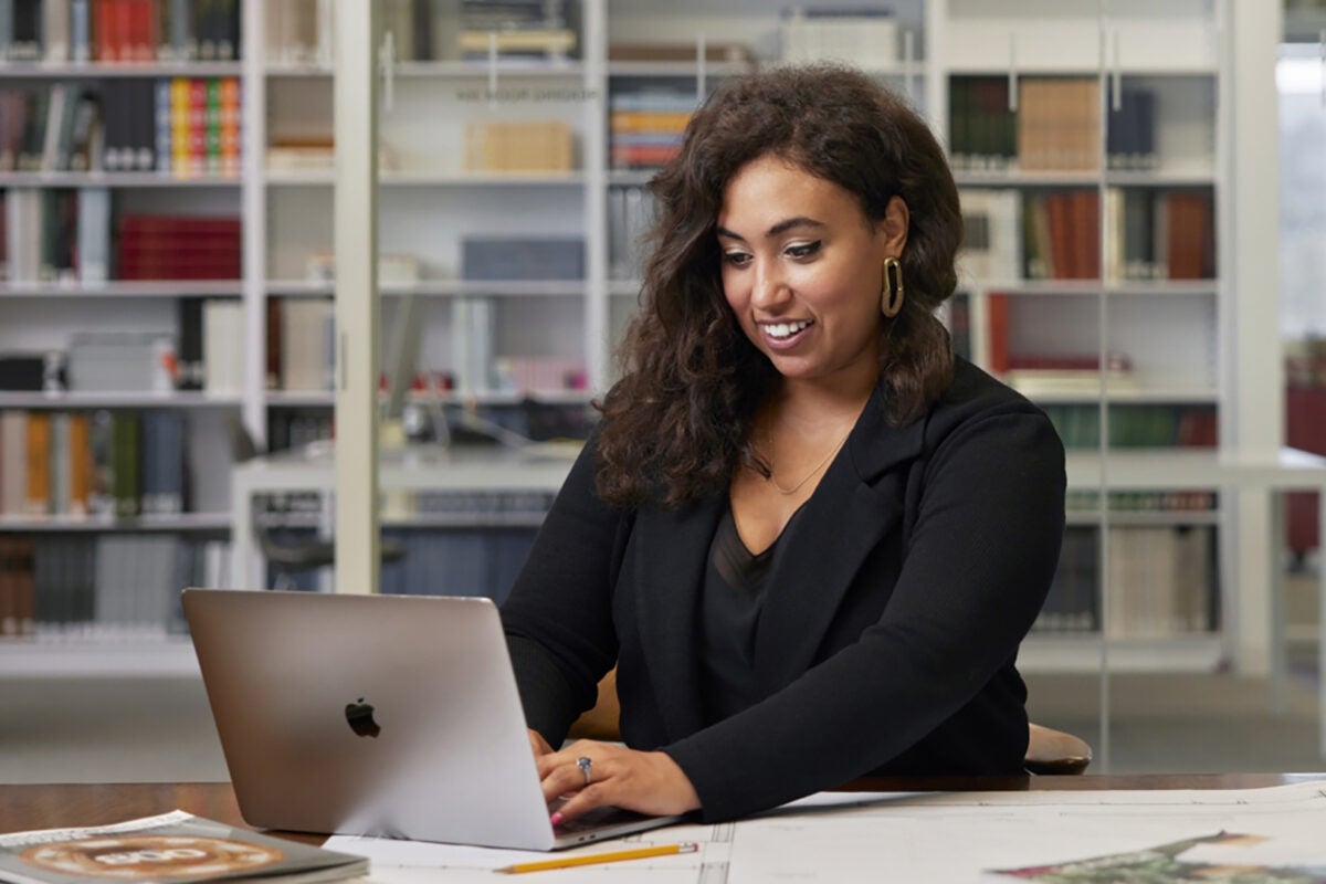 woman in a black suit working on a laptop with a bookshelf behind